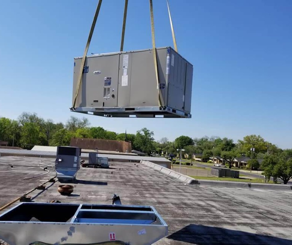 A large HVAC unit is being lifted by a crane to a rooftop under a clear blue sky, with nearby structures and a tree line visible. Installed by Wms Heating & Air Conditioning, a trusted AC Repair in Monroe, LA.