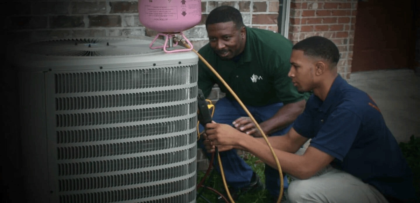 Two technicians are servicing an air conditioning unit outside a building, focusing on repairs with tools and equipment.