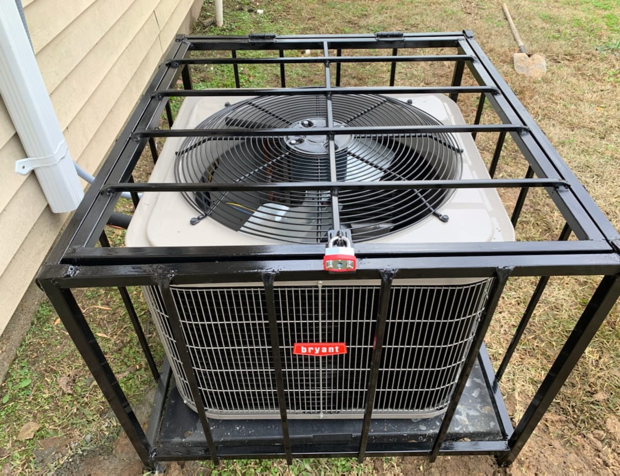 A metal cage surrounds a Bryant air conditioning unit, protecting it from potential damage. The unit is positioned beside a house on grass.